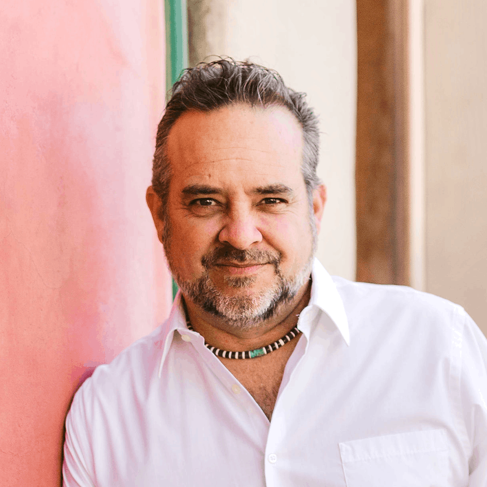 A middle-aged man with short dark hair and a beard, wearing a white shirt and beaded necklace, leans against a pink wall, smiling softly at the camera.
